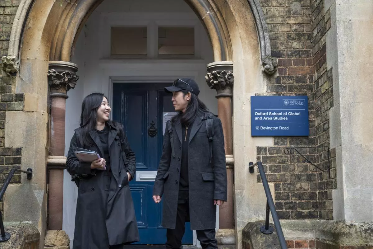 Two students standing outside the department front doors and arched porchway. They are wearing lightweight coats in dark colours.