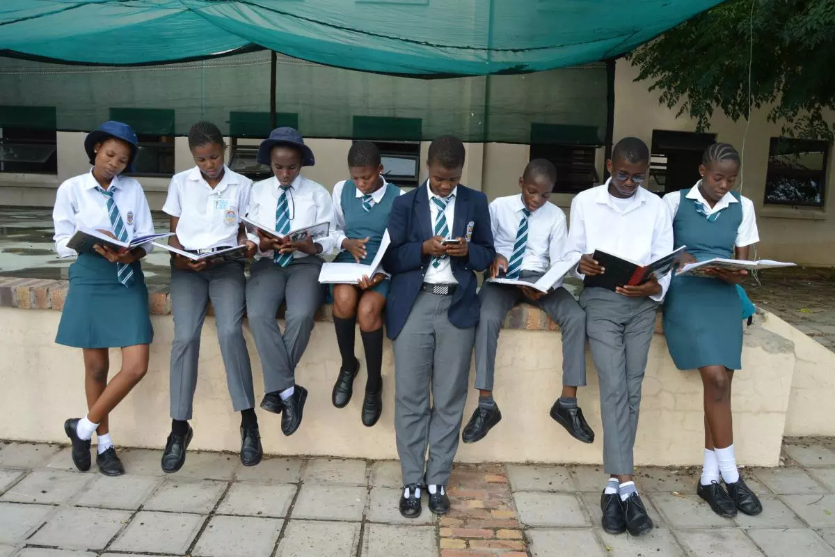 A row of school children in white and grey uniform sitting on a wall reading books. 