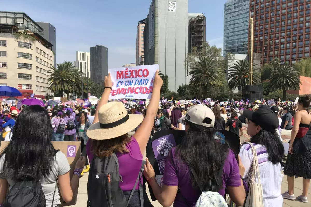 People walking along a street between high rise buildings. One is holding a sign 'Mexico es un Pais Feminicida' above her head. 