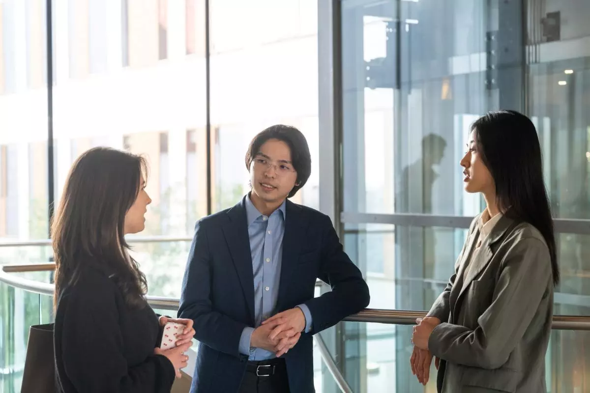 Three students standing in a corridor inside a modern glass building. Two are leaning on a railing and looking at the third student as they chat.