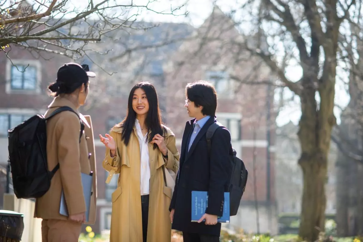 Three students standing outside chatting under a tree. 