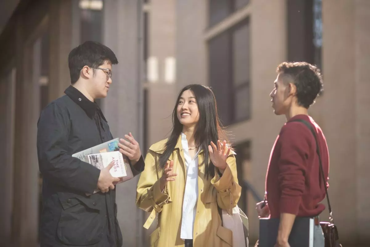 Three students standing outside chatting. They are wearing light jackets and two of them are holding books.