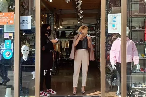 A lady dressed smartly in beige trousers, navy blue top and a jacket draped over her shoulders is leaving a clothes shop